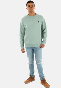 Sudadera verde claro con cuello redondo y logo, combinada con jeans azul claro y botas marrones. Textura suave, corte relajado.