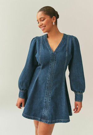 Femeie zâmbitoare, purtând o rochie albastră din denim cu mâneci lungi bufante și decolteu în formă de V, stând în fața unui fundal luminos și simplu.