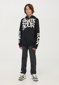 Felpa nera con grande grafica bianca "SKATE TOUR", tasca a marsupio e polsini a coste. Accoppiata con jeans grigio scuro e sneakers bianco e nere.