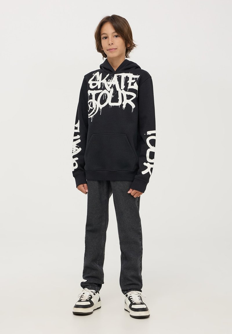 Felpa nera con grande grafica bianca "SKATE TOUR", tasca a marsupio e polsini a coste. Accoppiata con jeans grigio scuro e sneakers bianco e nere.