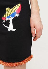 Svart kjol med en fransad nederdel i orange och gult. Har en illustration av en vit katt som bär en stor halmhatt och solglasögon.