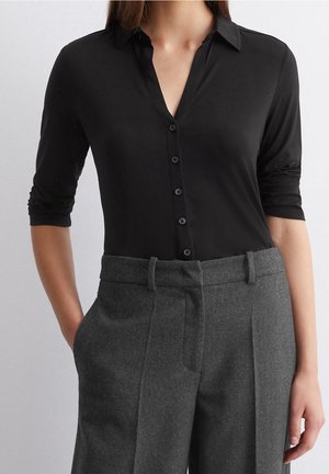 Kobieta ubrana w czarną, zapinaną na guziki koszulę z podwiniętymi rękawami, wpuszczoną w wysokiej talii szare wełniane spodnie z szlufkami na pasek.