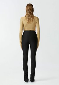 Bodysuit dugih rukava bež boje s glatkom teksturom, uparen s crnim tajicama visokog struka. Oba komada imaju pripijen dizajn.