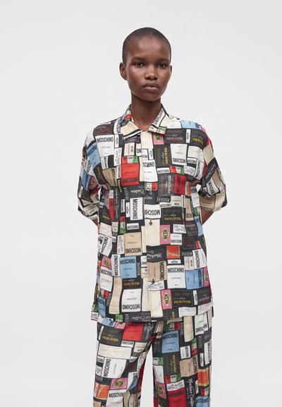 Personne portant une chemise et un pantalon assortis couverts d'imprimés colorés de la marque de mode Moschino sur un fond blanc uni.