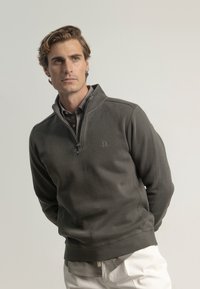 Suéter gris con cuello de media cremallera, textura acanalada y logo sutil. Llevado sobre una camisa con cuello y combinado con pantalones blancos.