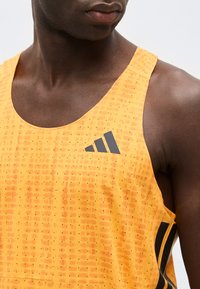 Κοντινό πλάνο ενός ατόμου που φοράει πορτοκαλί αθλητική μπλούζα χωρίς μανίκια με υφή και μαύρο λογότυπο Adidas στο στήθος.