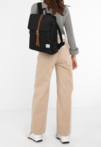 Femme portant un pantalon beige et un pull gris porte un sac à dos noir avec une sangle marron et une étiquette blanche dans le dos.