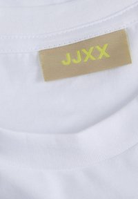 Bílý tričko s kulatým výstřihem, texturovaná látka, se béžovou nášivkou s nápisem "JJXX" v jasně žlutých písmenech přišitou na vnitřním límci.