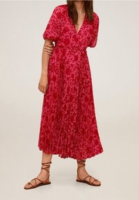 Robe midi à fleurs rouges avec des manches courtes bouffantes, décolleté en V et jupe plissée. Associée à des sandales à lanières marron.