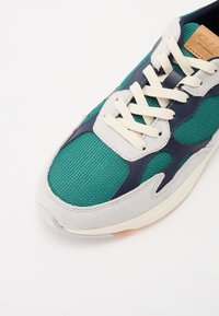 Sneakers in rete verdi con accenti in suede grigio e blu navy. Presentano lacci beige e una tomaia testurizzata. Suola in gomma leggera con una leggera trazione.