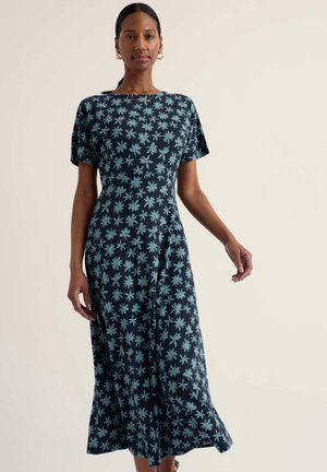 Femeie purtând o rochie midi bleumarin cu model floral albastru deschis, mâneci scurte și cercei de tip cerc auriu, stând pe un fundal simplu.