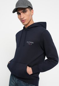 Námornícky modré mikina s kapucňou, ktorá má predné vrecko, kapucňu so šnúrkou a vyšívané logo "HILFIGER" s rokom 1985 v bielej a červenej farbe.