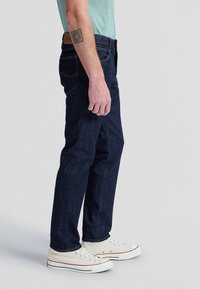 Homme portant un jean bleu foncé et des baskets en toile blanches, debout de profil devant un fond uni.