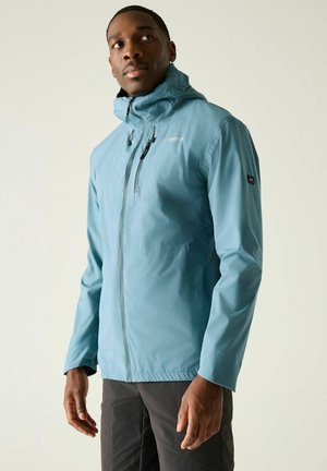 Man wearing light blue zip-up hooded jacket and dark pants, looking slightly to the side against a plain background.