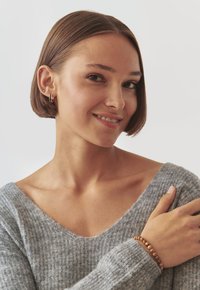 Suéter gris de punto con escote en V. Pulsera de cuentas de madera marrón en la muñeca, aretes de aro dorados. Cabello liso y textura de piel suave.