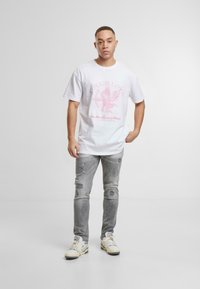 Camiseta blanca de algodón con un diseño gráfico rosa que presenta un ángel, combinada con jeans grises desgastados y zapatillas beige.