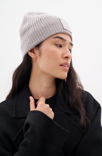 Světle šedý žebrovaný pletený čepice s ohrnutým lemem, se malou obdélníkovou nášivkou. Model má na sobě černý kabát, ruku položenou na límci.