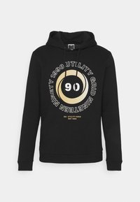 Sudadera negra de tela suave, con un diseño gráfico circular con detalles en dorado y blanco, que incluye los números "90" y "1990."