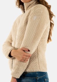 Giacca in pile beige, tattile, con colletto alto e chiusura con zip. Presenta un logo sulla manica sinistra e un tessuto morbido e accogliente.
