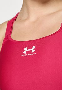 Sujetador deportivo rojo fabricado en tejido suave, con escote redondeado, tirantes finos y el logo blanco de Under Armour en la parte frontal.