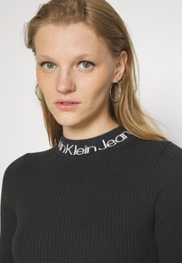 Zwarte geribbelde gebreide top met een hoge kraag en witte "Calvin Klein Jeans" tekst. Model draagt grote oorbellen. Geverfd stof en aansluitend ontwerp.