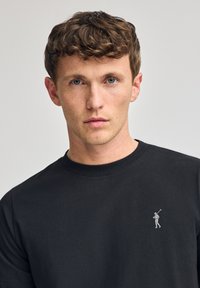 T-shirt in cotone nero con collo a giro, caratterizzato da un piccolo logo argentato di un golfista sul lato sinistro del petto. Texture liscia, design casual.