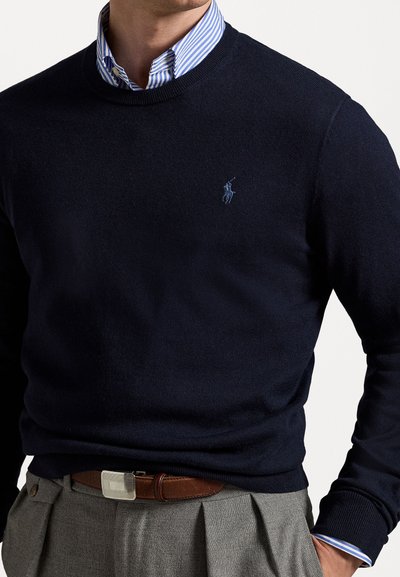 Suéter de punto azul marino con cuello redondo y un pequeño logo azul. Usado sobre una camisa de cuello blanco y azul a rayas con puños visibles.