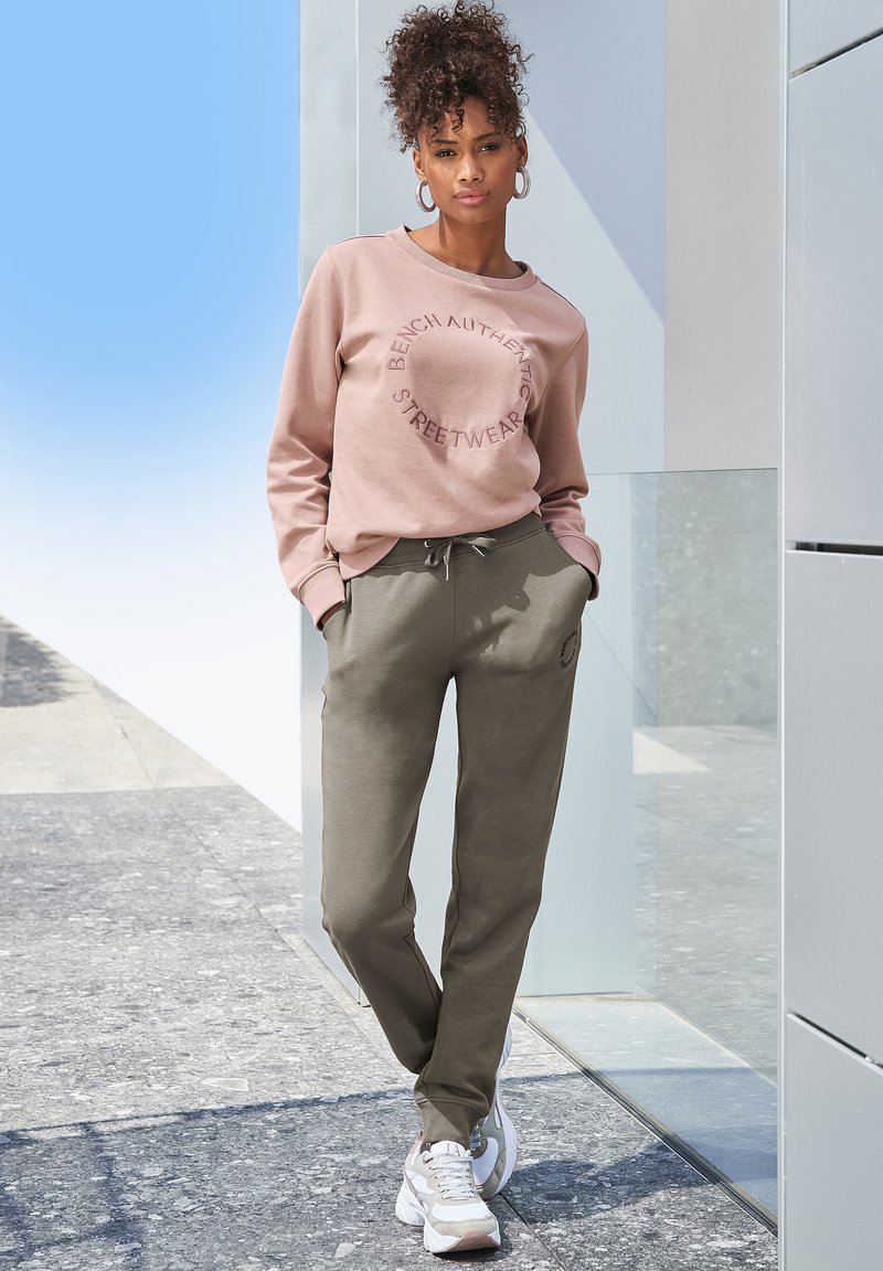 Lichtroze sweatshirt met een ronde logo, gecombineerd met donkergroene joggingsbroek met een trekkoord. Khaki sneakers maken de outfit compleet.