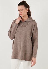 Brauner Strickpullover mit einem Reißverschlusskragen, geripptem Detail am Hals, lässiger Passform und langen Ärmeln, getragen über weißen Hosen.