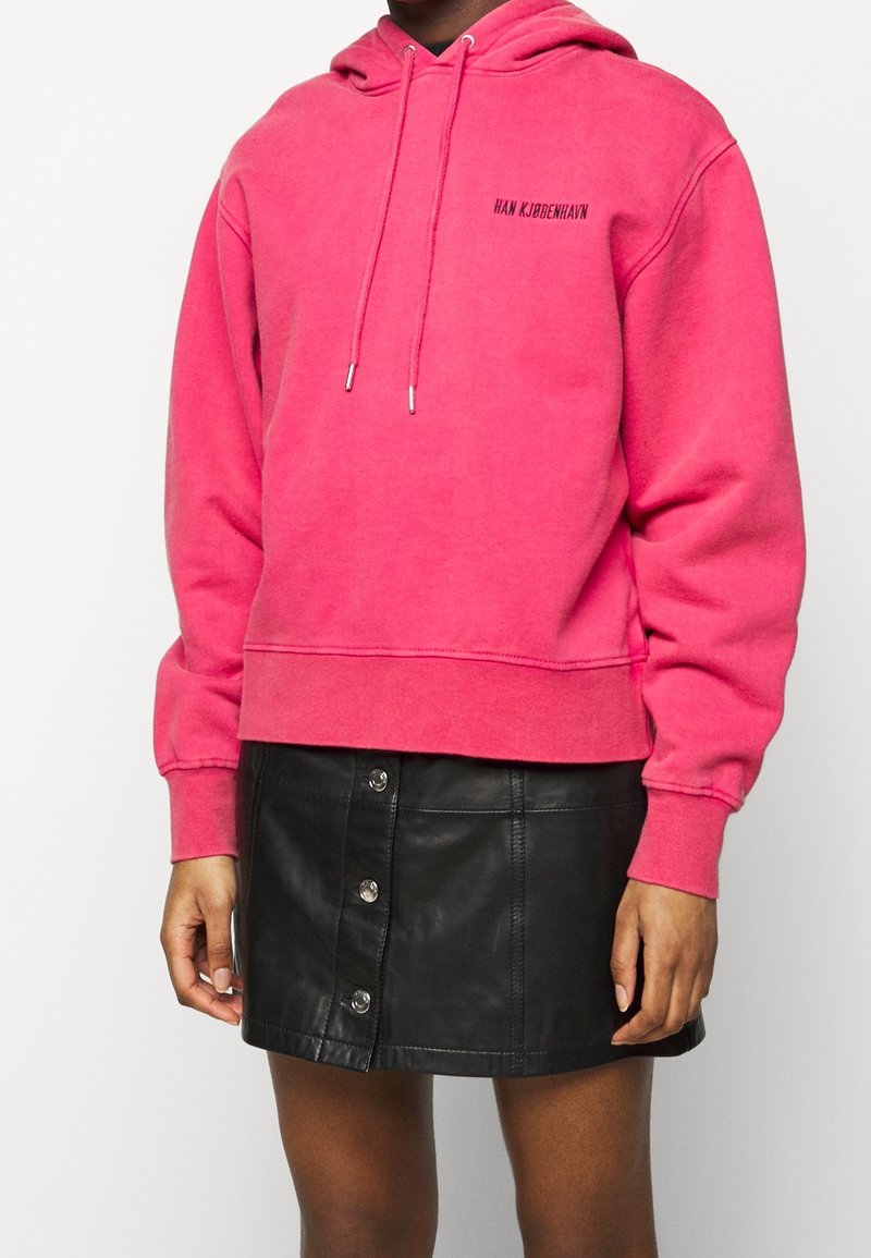Personne portant un sweat à capuche rose vif avec des cordons de serrage et un petit texte noir, associée à une jupe en cuir noire boutonnée sur le devant.