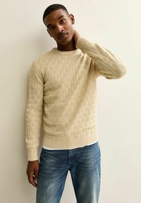 Beige reliëftrui met een kabelbreipatroon, ronde hals en geribde manchetten. Draagt over een wit shirt, gecombineerd met blauwe jeans.