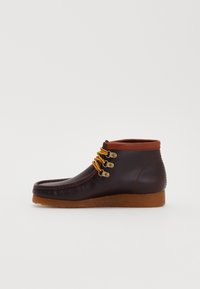 Botas de cuero marrón hasta el tobillo con punta redondeada, cuello naranja contrastante, cordones amarillos y tres ojales dorados. Suela de goma.