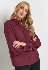 Blusa borgogna con colletto alto, tessuto testurizzato, maniche lunghe e polsini con bottoni. La silhouette è ampia e leggermente corta.
