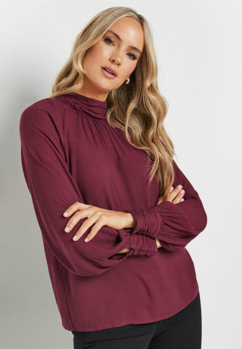 Blusa borgogna con colletto alto, tessuto testurizzato, maniche lunghe e polsini con bottoni. La silhouette è ampia e leggermente corta.
