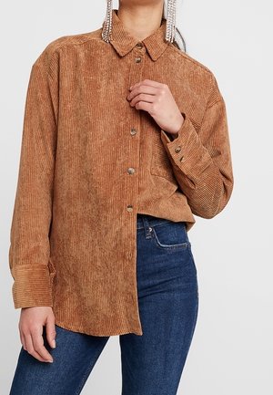 Femme portant une chemise en velours côtelé marron à boutons avec manches longues et un jean en denim bleu sur un fond uni.
