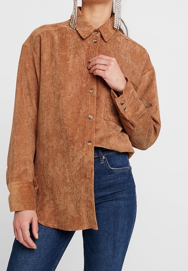 Femme portant une chemise en velours côtelé marron à boutons avec manches longues et un jean en denim bleu sur un fond uni.