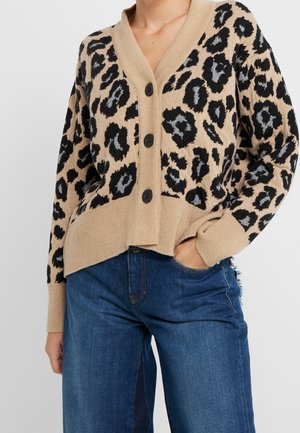Femme portant un cardigan beige boutonné avec un imprimé léopard noir et gris et un jean bleu taille haute, une main dans la poche.