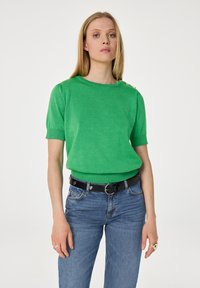 Blonde vrouw die een felgroene korte mouwen trui met gouden knopen op de schouders draagt, blauwe jeans en een zwarte riem met hartvormige details.