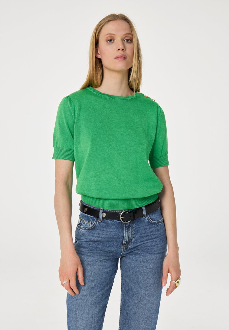 Blonde vrouw die een felgroene korte mouwen trui met gouden knopen op de schouders draagt, blauwe jeans en een zwarte riem met hartvormige details.