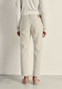 Jeans en denim beige clair avec une coupe ajustée, dotés de deux poches arrière et d'une pièce en cuir marron sur la taille. Portés avec des tongs.