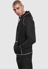 Crna majica s kapuljačom i vezicama, sa reflektirajućim šavovima duž šavova i prednjim džepom za dodatnu funkcionalnost.