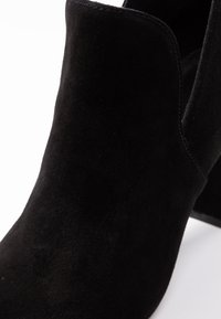 Botín de ante negro con una textura suave, punta redonda y un tacón bajo y ancho. La abertura tiene un diseño de bordes suaves, ofreciendo una silueta elegante.