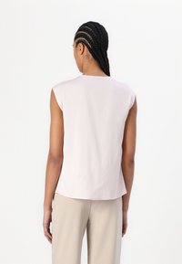 Top senza maniche rosa chiaro in tessuto morbido, con scollatura rotonda e orlo leggermente curvato. Indossato con pantaloni beige.