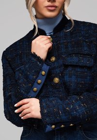 Mörkblå tweedjacka med guldfärgade knappar, texturerad tyg och fransade kanter. Har en krage och en ficka på vänster sida.