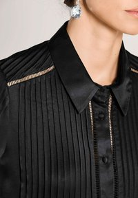 Zwarte satijnen blouse met verticale plooien, een klassieke kraag, knoopsluiting en doorschijnende paneelaccenten langs de schouders.