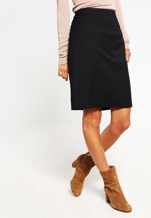 Femme portant une jupe noire mi-longue, un haut à manches longues rose clair et des bottines marron en suède à talons, debout jambes croisées.