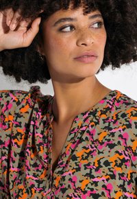 Patroond blouse met een mix van roze, oranje en zwarte vormen op een beige achtergrond. V-hals, zachte stof en een losse pasvorm.
