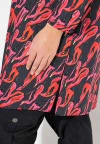Camicia colorata con un design fluente, caratterizzata da motivi ondulati rossi e rosa su uno sfondo nero, abbinata a pantaloni scuri e con dettagli di bottoni.