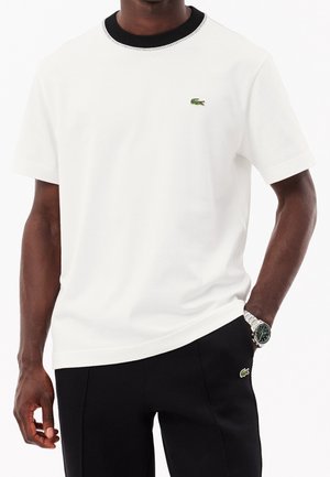 T-shirt blanc à manches courtes, avec un col côtelé noir et un logo vert brodé sur la poitrine. Texture lisse et coupe décontractée.