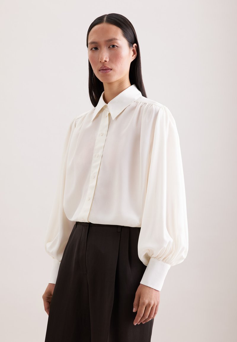 Femme aux longs cheveux foncés portant un chemisier blanc à manches bouffantes et un pantalon noir taille haute, debout devant un fond clair uni.